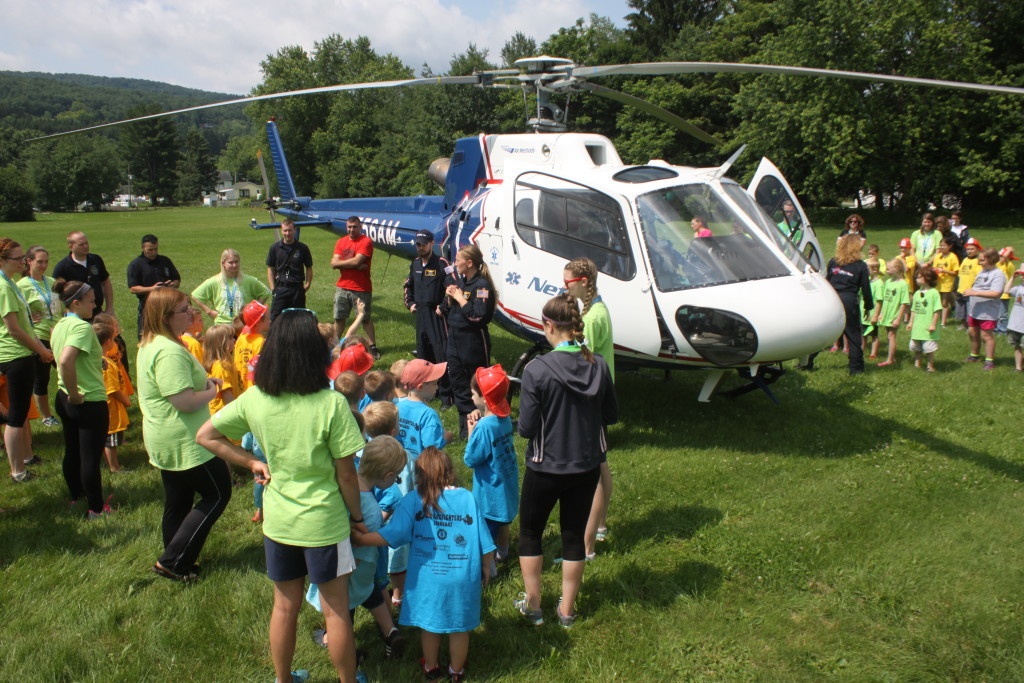 Over 60 children graduated from the YMCA Jr. Firefighters program this afternoon, where, after learning fire safety, they received medals of completion, and got an up-close and personal look at the Life Net Helicopter which flew in from Sidney. Pilot Lindsey Stone, center, talked to the kids about the specs of the of the helicopter and safety equipment like helmets and seat belts. (Ian Austin/allotsego.com)