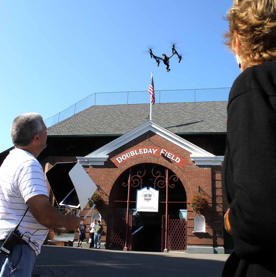 Paul Frederick of Adworkshop, Lake Placid, launches a drone over Doubleday Field a few minutes ago, which is shooting footage of "The Birthplace of Baseball" in preparing new web and social media videos for Destination Marketing Corp. of Otsego County, the county's tourism promotion effort. DMOC's director, Deb Taylor, left, said the Adworkshop team will be in the county through Thursday; the drone will be buzzing over Hyde Hall tomorrow, and other shoots are still being arranged. (Jim Kevlin/allotsego.com)