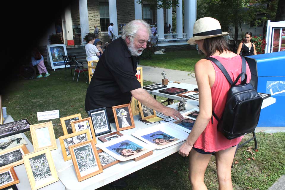 Joseph Kurhajec of Paris, Merida, Mexico, and Treadwell, Delaware County, answers questions from Chelsea Guss, New York City, who is visiting her family’s summer home in Middlefield this weekend, at The Cooperstown Art Association’s Fine Art on the Lawn, underway at 22 Main until 5 this evening and 10-5 Sunday. Kurhajec is a sculptor who has spent part of the year in Delaware County since 1971, where he operates the Treadwell Museum of Fine Art. He also spends part of each year at homes in France and Mexico. (Jim Kevlin/allotsego.com)