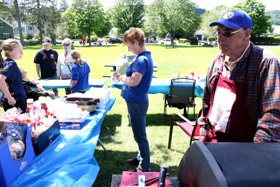 Rotarian Chuck Newman mans the grill, and Diana Nicols (assisted by daughters Tess, left, and Tara) take orders for dogs and burgers at the club's stand in Cooperstown's Lakefront Park at this hour.  Business will pick up after 5, as Fourth of July celebrants begin arriving in force, and after 7 in particular.  Fireworks start at dusk, which today happens at 9:16 p.m.  (Jim Kevlin/AllOTSEGO.com)