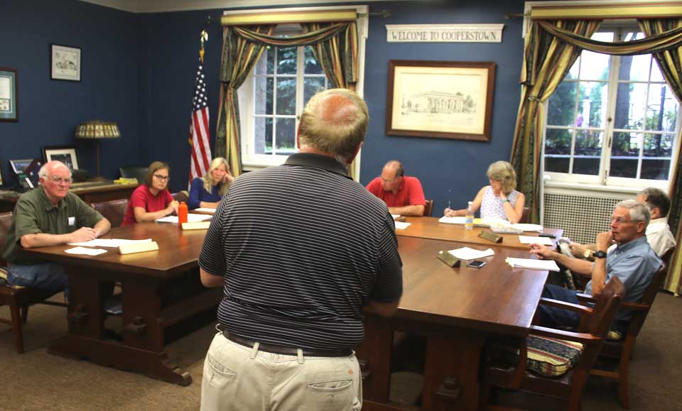 Vin Russo, Mickey’s Place proprietor, (back to camera), suggests to the Cooperstown Village Board at this evening meeting that it form an “entity” to interface with the Hall of Fame over issues that rose during this Induction Weekend, then to do so again in advance of future Inductions. “There were a lot of things that did come up,” said Russo. “While it is fresh in our minds, I think we should discuss what we might do about it.” One current issue is the metal fences the Hall installs along Main Street, how close they should be to the curbs and whether there are sufficient breaks for people to cross the street. Further, Russo said, often people don’t know where to raise questions, with Village Hall, the Baseball Hall, police or whatever. After he departed, Trustee Cindy Falk said, “I think Vinnie’s idea is a good one.” Mayor Jeff Katz assigned the question to Falk’s Economic Development Committee, with perhaps forming such an entity during August. Seated, from left, are Trustees Jim Dean, Falk and Ellen Tillapaugh Kuch, Village Attorney Martin Tillapaugh, Mayor Katz, Village Clerk Teri Barown, and Trustees Richard Sternberg and Lou Allstadt. (Jim Kevlin/AllOTSEGO.com)