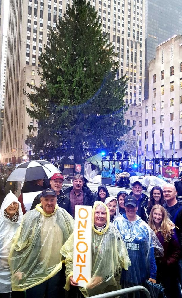 A contingent from Oneonta arrives at Rockefeller Plaza a few minutes ago, looking forward to the lighting of the Eichler family’s 95-foot-tall blue spruce at 8:55 this evening. From left are Linda Rae-Nichols, Ted Gaisford, Darren Gaisford, Todd Foreman, Emmy Gaisford (with sign), Sue Gillette, June Sheehan, Tracy Gaisford, Greg Krikorian and his two daughters, Bridget and Claire, and Brian Gillette. The contingent rode one of Eastern Travel’s chartered buses from the City of the Hills to the Big Apple today. (Ian Austin/AllOTSEGO.com)